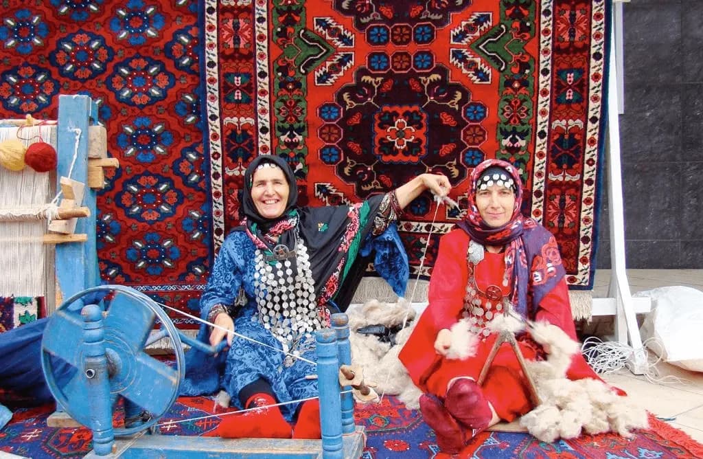 Две мастерицы в национальных костюмах прядут шерсть на фоне развешанных дагестанских ковров