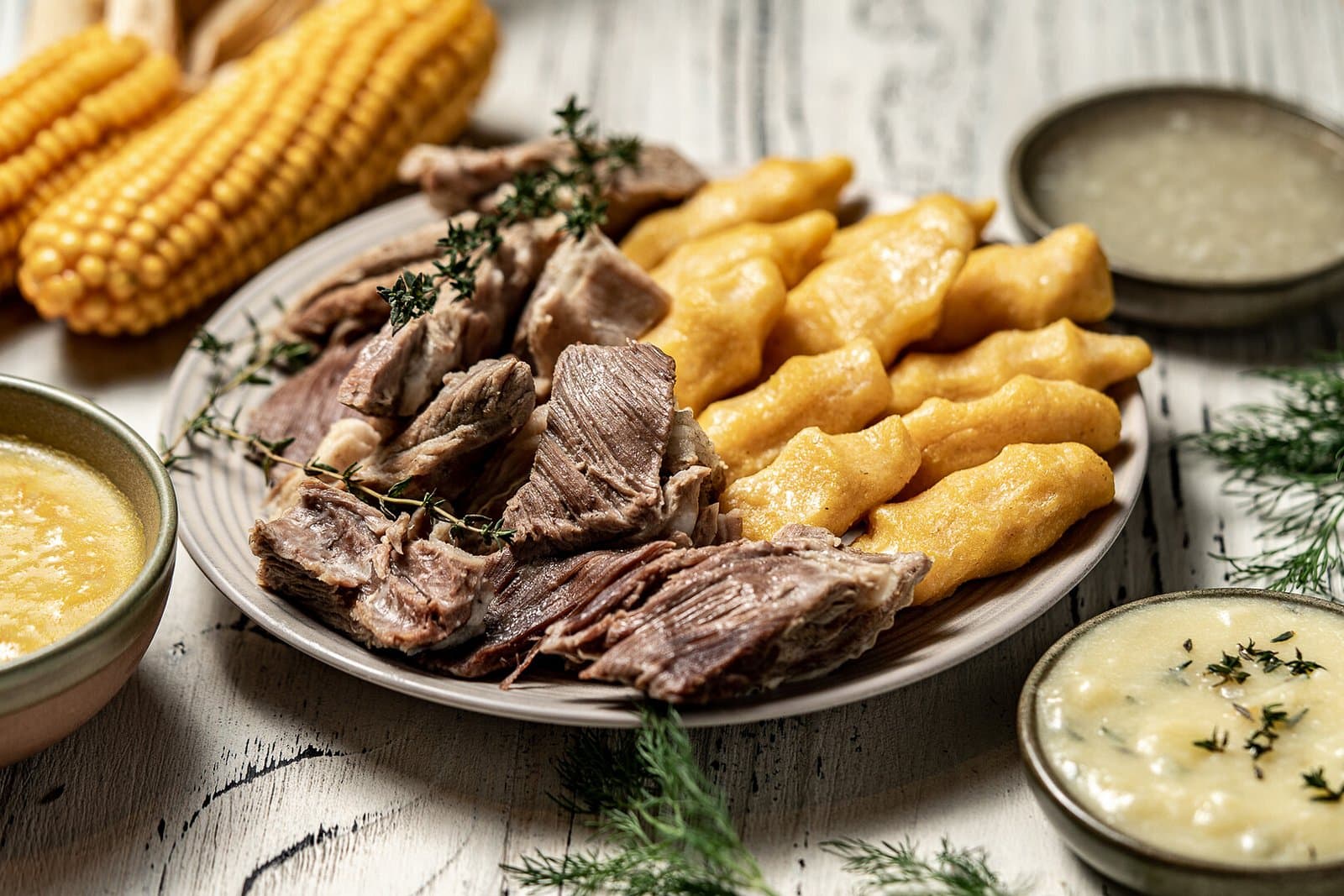 Кусок вяленого мяса и продолговатые кукурузные галушки халтама на тарелке, рядом початки кукурузы и две мисочки соуса