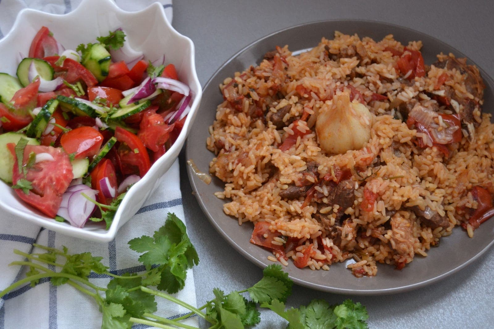 Лезгинский плов на плоской тарелке: рис с томатом и кусочками мяса, в центре целая головка чеснока, рядом салат из огурцов и помидоров