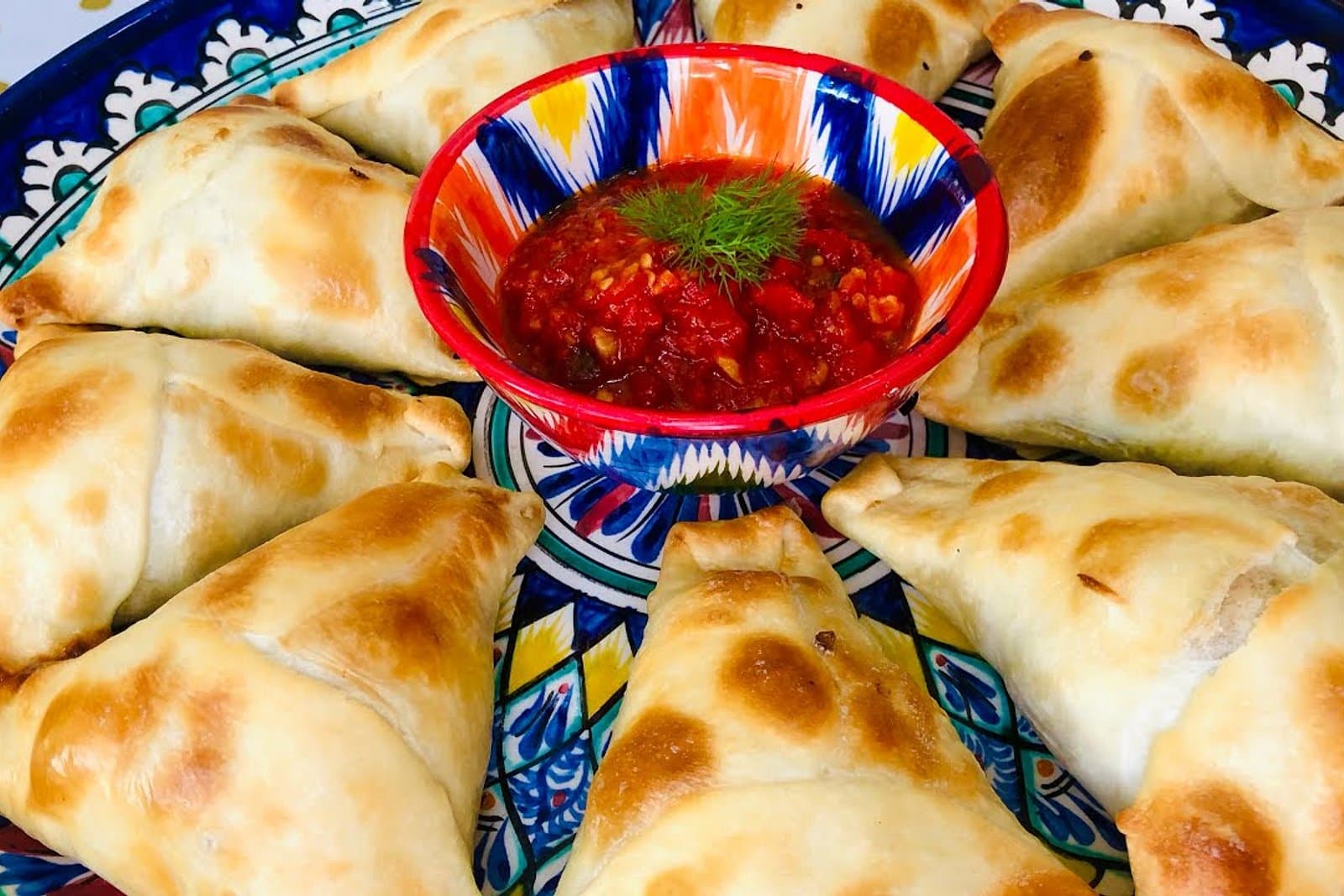 Треугольные самсы с кунжутом, выложенные кругом на узорной тарелке вокруг мисочки с томатным соусом и укропом