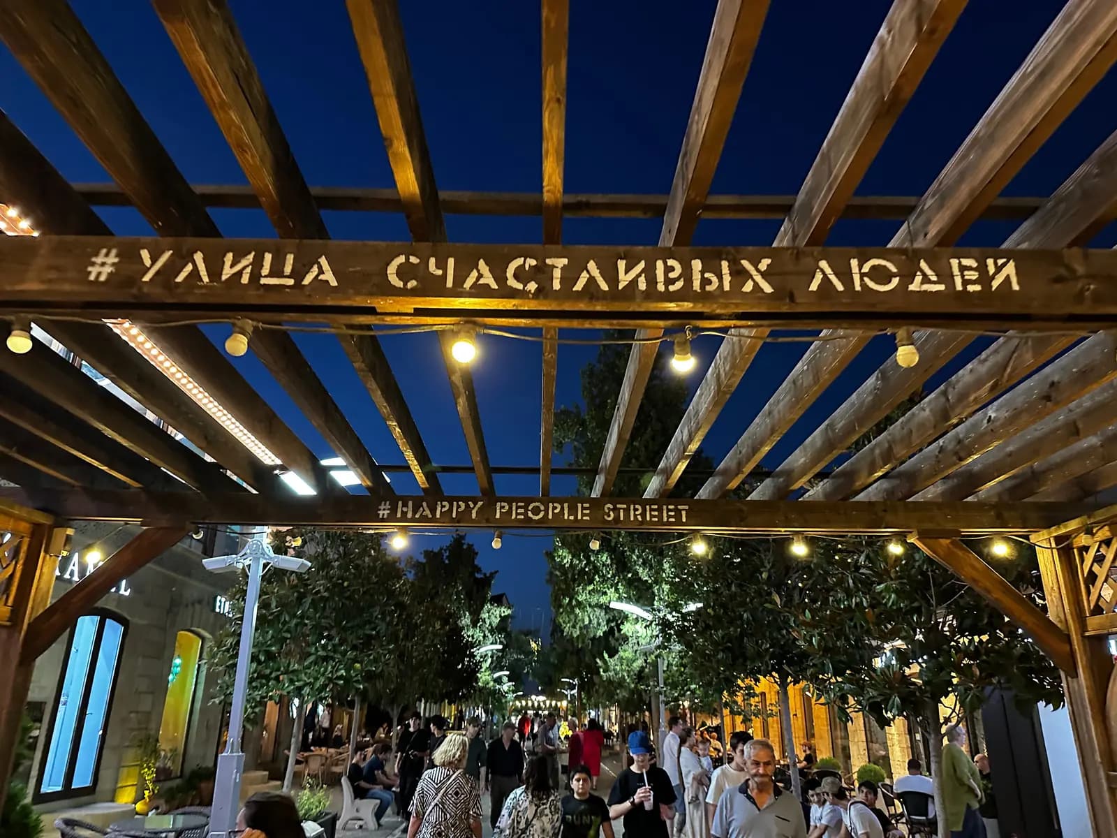 Деревянная пергола с надписью «Улица счастливых людей» и гирляндами над пешеходной улицей в Дербенте вечером