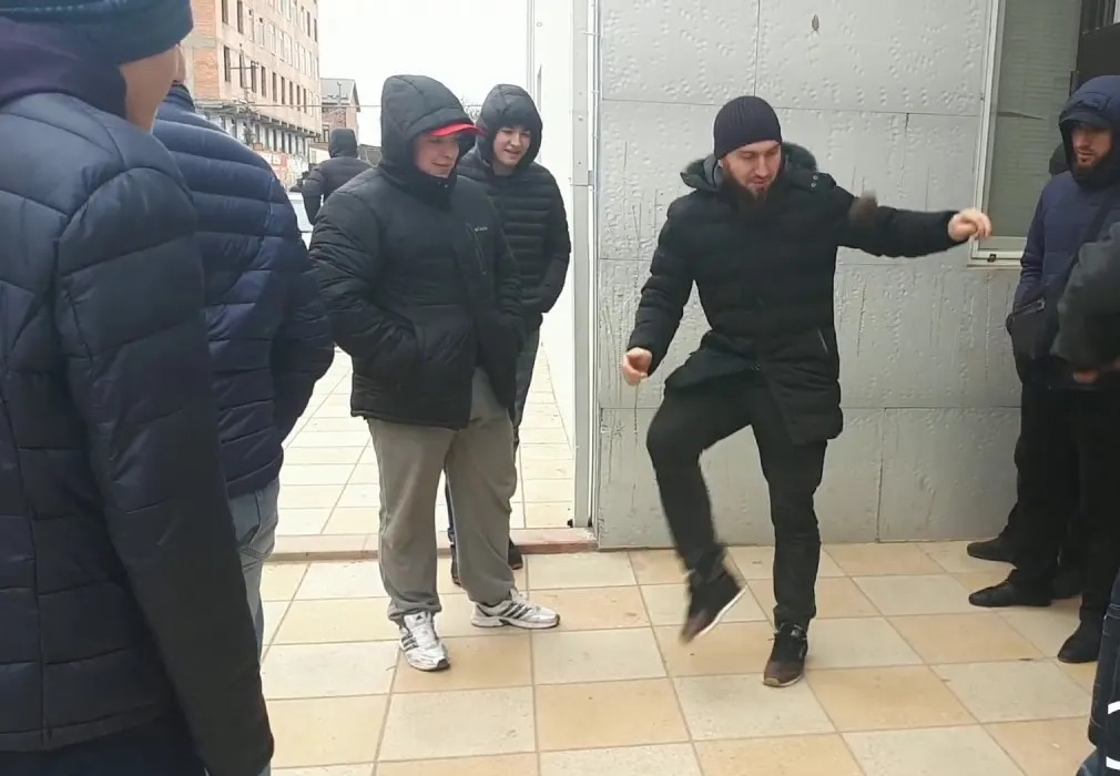 Мужчина в зимней куртке подбрасывает лянгу ногой, вокруг во дворе стоят зрители