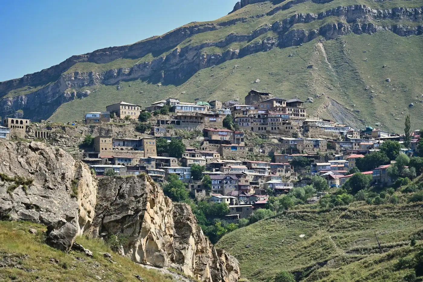 Панорама аула Чох в Дагестане летом — жёлтая каменная застройка на скалистом склоне среди зелёных гор
